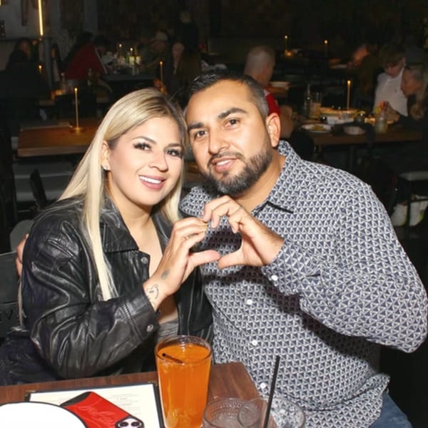 A smiling couple forms a heart with their hands at a dimly lit restaurant table.