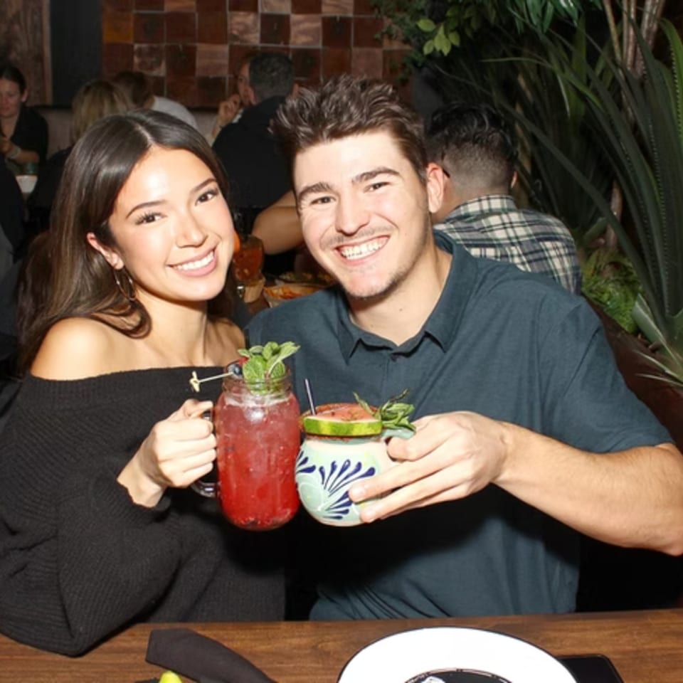 Smiling couple holding cocktails at a restaurant table with plants in the background.