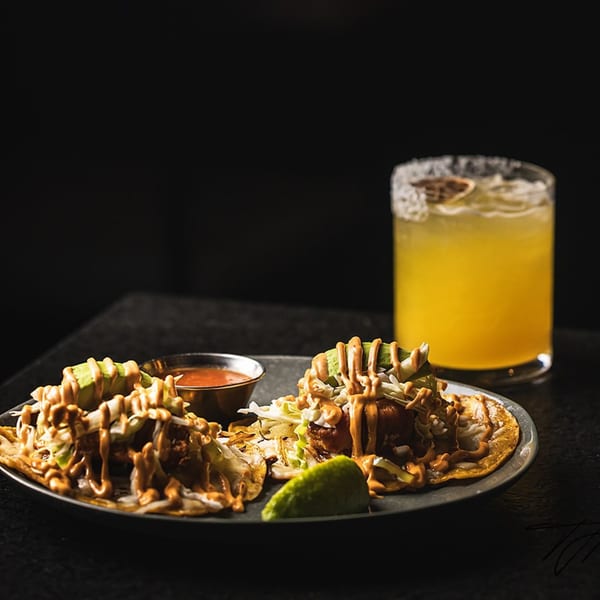 Two tacos with sauce, avocado, and cabbage, with a margarita and salsa on a dark table.
