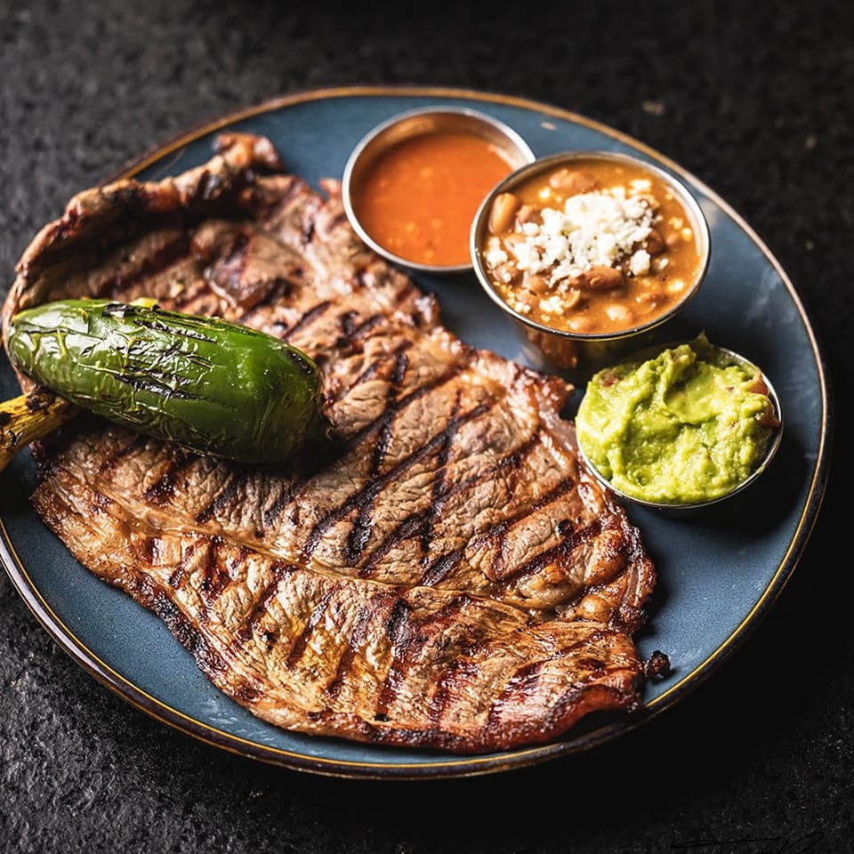 Grilled steak with a charred jalapeño, guacamole, beans, and red sauce on a blue plate.