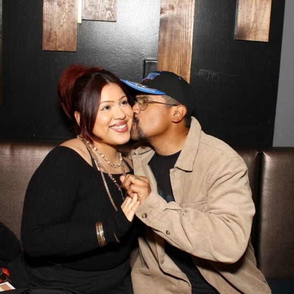 A man in a cap kisses a smiling woman on the cheek while holding her hand in a restaurant booth.