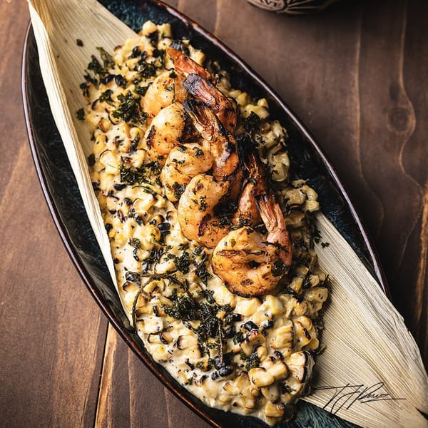 Grilled shrimp on a bed of creamy corn and dark herbs, served in a corn husk.