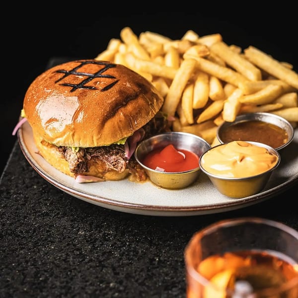 Pulled pork burger with fries and three dipping sauces on a plate, with a drink in the foreground.