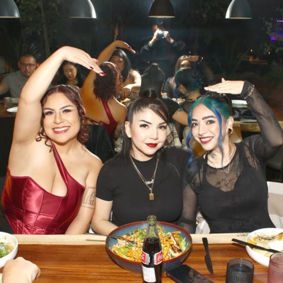 Three women posing with hands on heads at a restaurant table with food and drinks.
