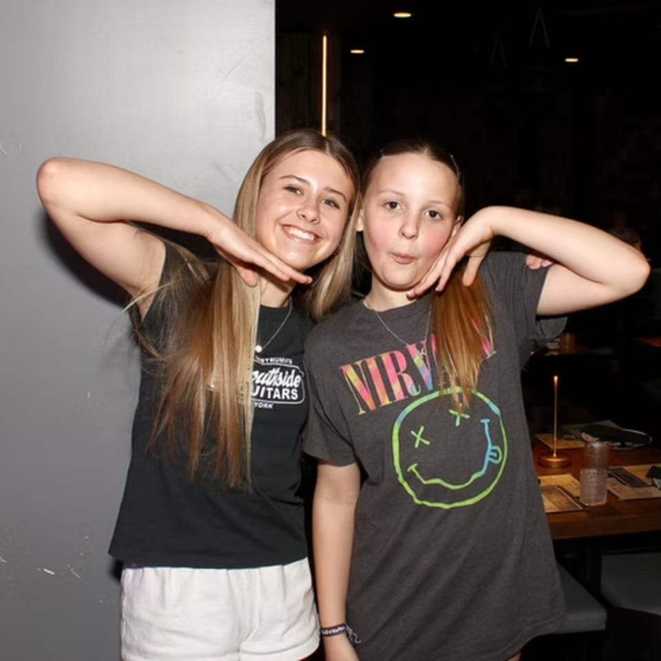 Two young girls pose playfully, smiling at the camera in a dimly lit restaurant.