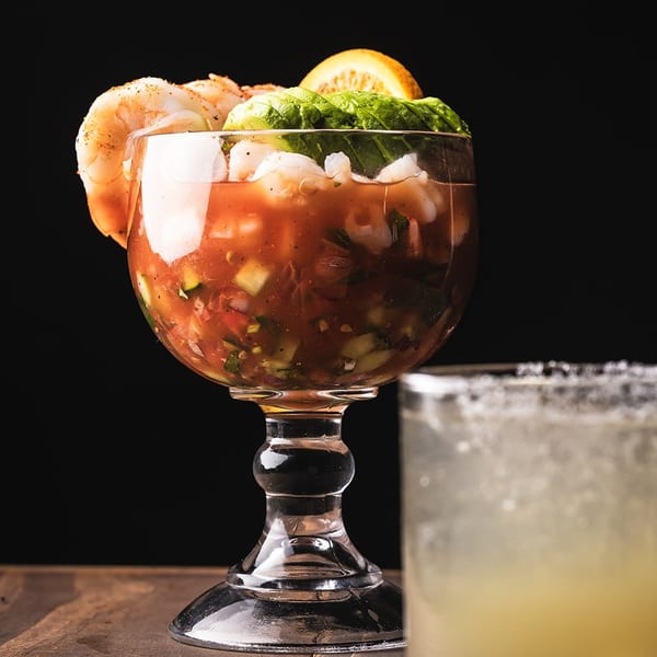 Shrimp cocktail in a goblet with avocado and orange, next to a margarita.