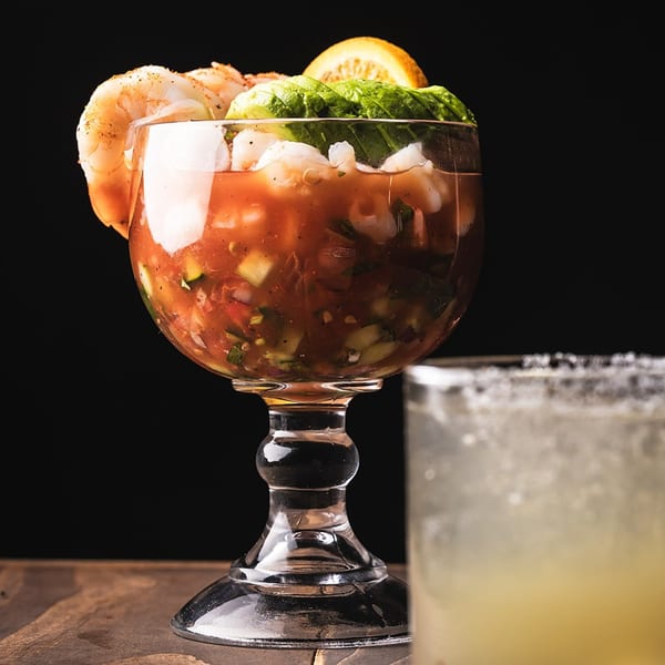 Shrimp cocktail with avocado and orange slices, next to a margarita on a wooden table.