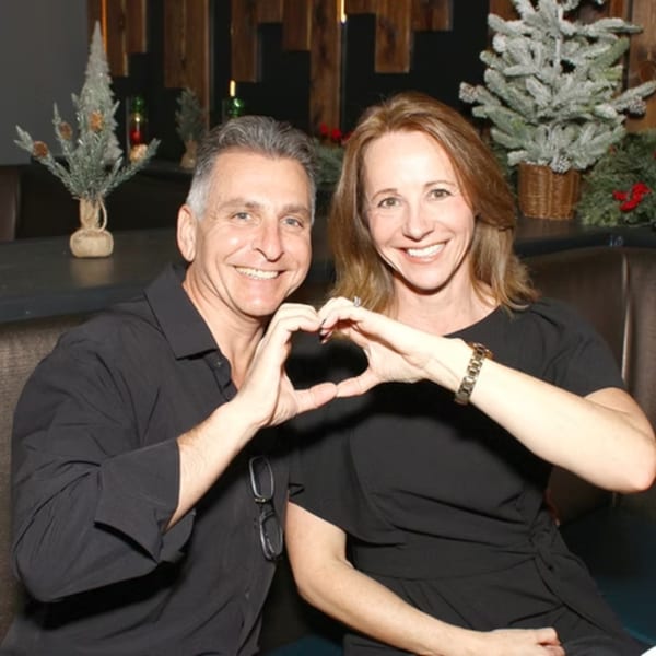 Smiling couple in black shirts making a heart shape with their hands, with holiday decorations in the background.
