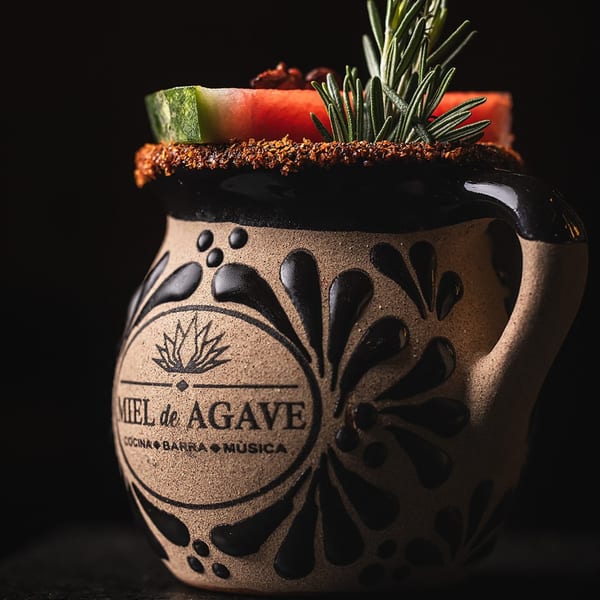 Miel de Agave branded mug with a drink garnished with watermelon, rosemary, and a spiced rim.