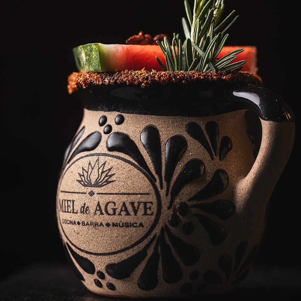 Miel de Agave branded mug with a drink garnished with watermelon, rosemary, and a spiced rim.