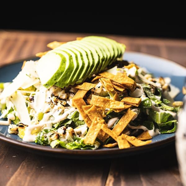 A vibrant salad with sliced avocado, tortilla strips, charred corn, and creamy dressing on a blue plate.