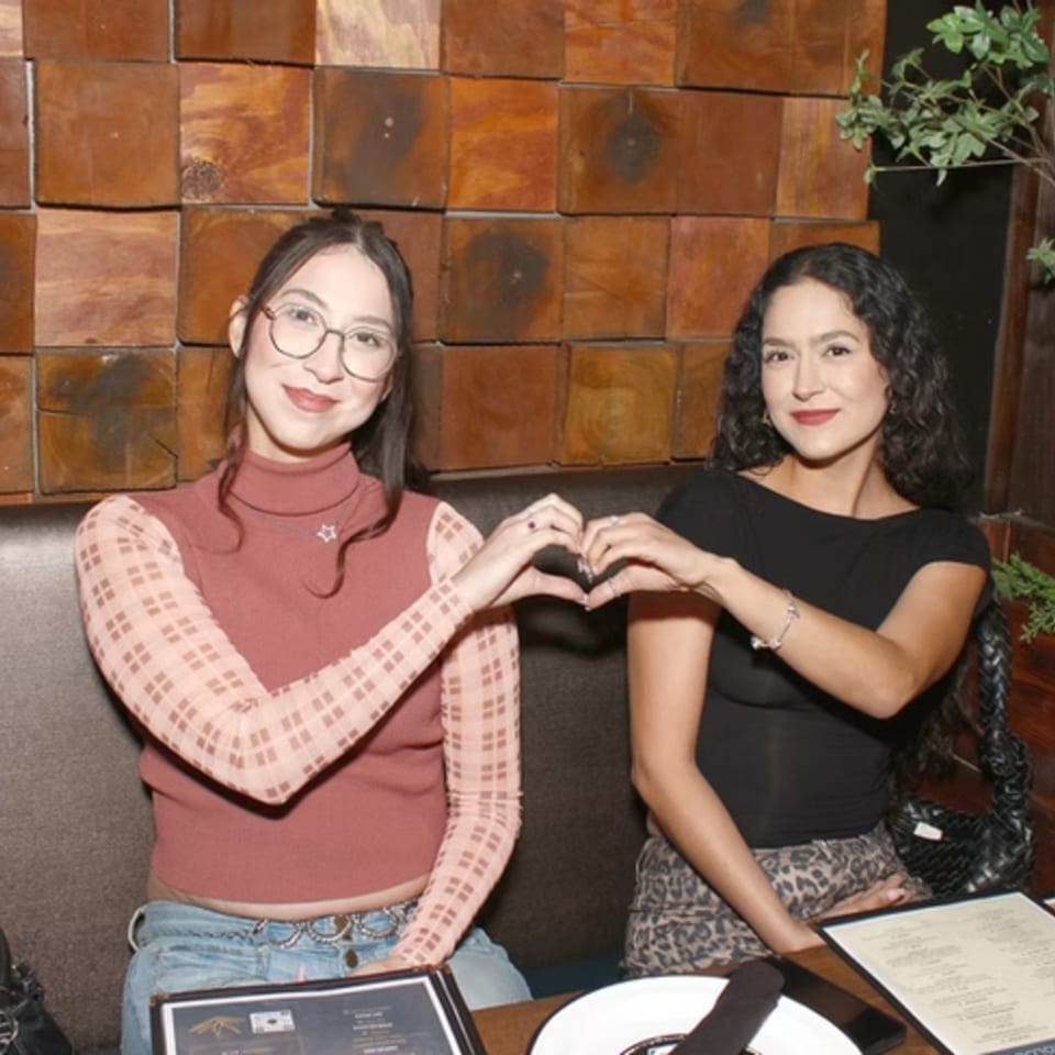 Two women sitting at a restaurant table, making a heart shape with their hands.