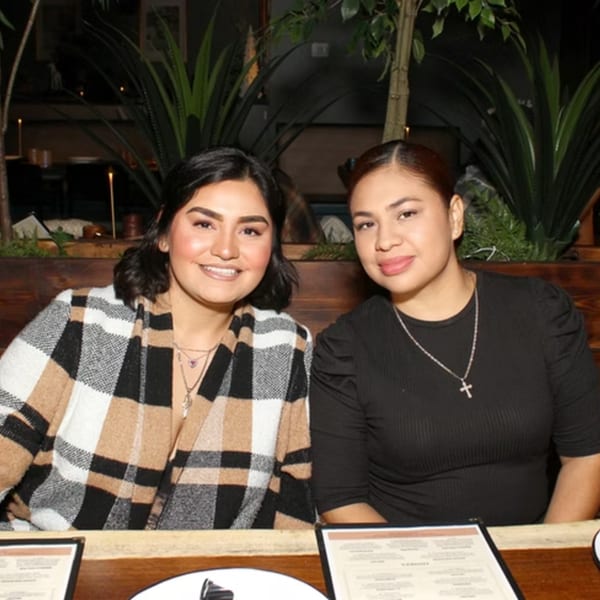 Two smiling women seated at a restaurant table with menus, surrounded by plants and warm lighting.