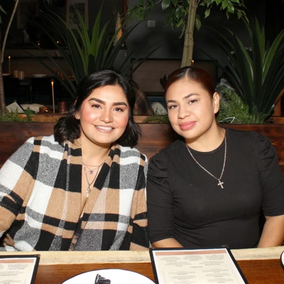 Two smiling women seated at a restaurant table with menus, surrounded by plants and warm lighting.