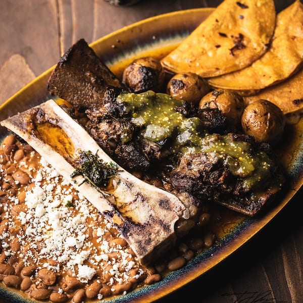 Hearty Mexican dish with short ribs, bone marrow, beans, potatoes, and tortillas on a rustic plate.
