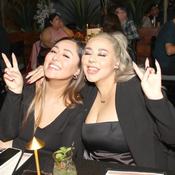Two smiling women pose for a photo at a restaurant table with drinks and other patrons in the background.
