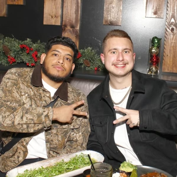 Two men smiling and making peace signs at a restaurant table with food and drinks.