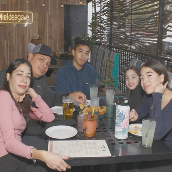 A family of five smiles at the camera while seated at a restaurant table with drinks and food.