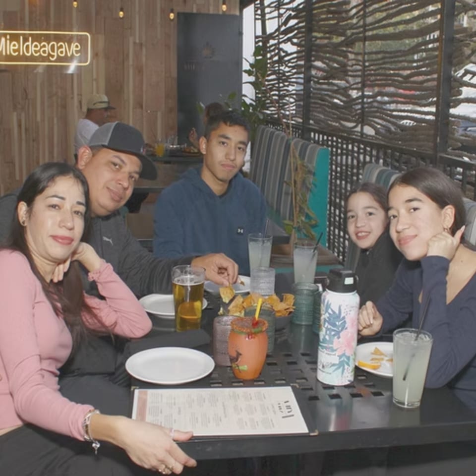 A family of five smiles at the camera while seated at a restaurant table with drinks and food.