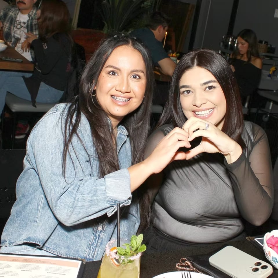 Two smiling women making a heart shape with their hands at a restaurant table.