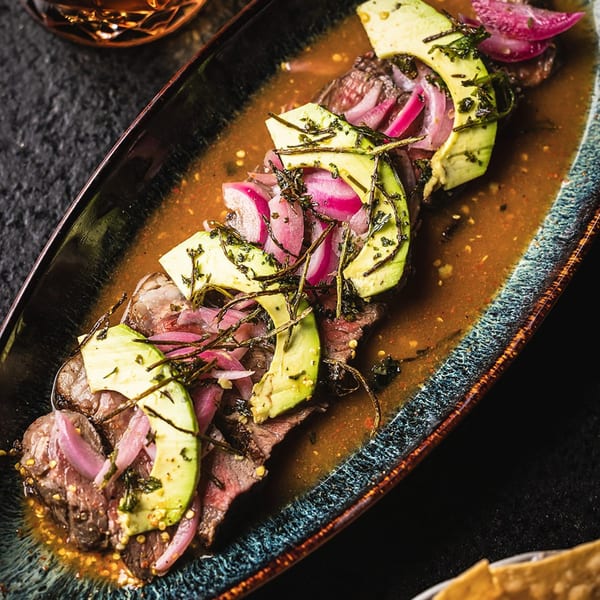 Sliced steak with avocado, pickled red onions, and herbs in a savory sauce on a dark platter.