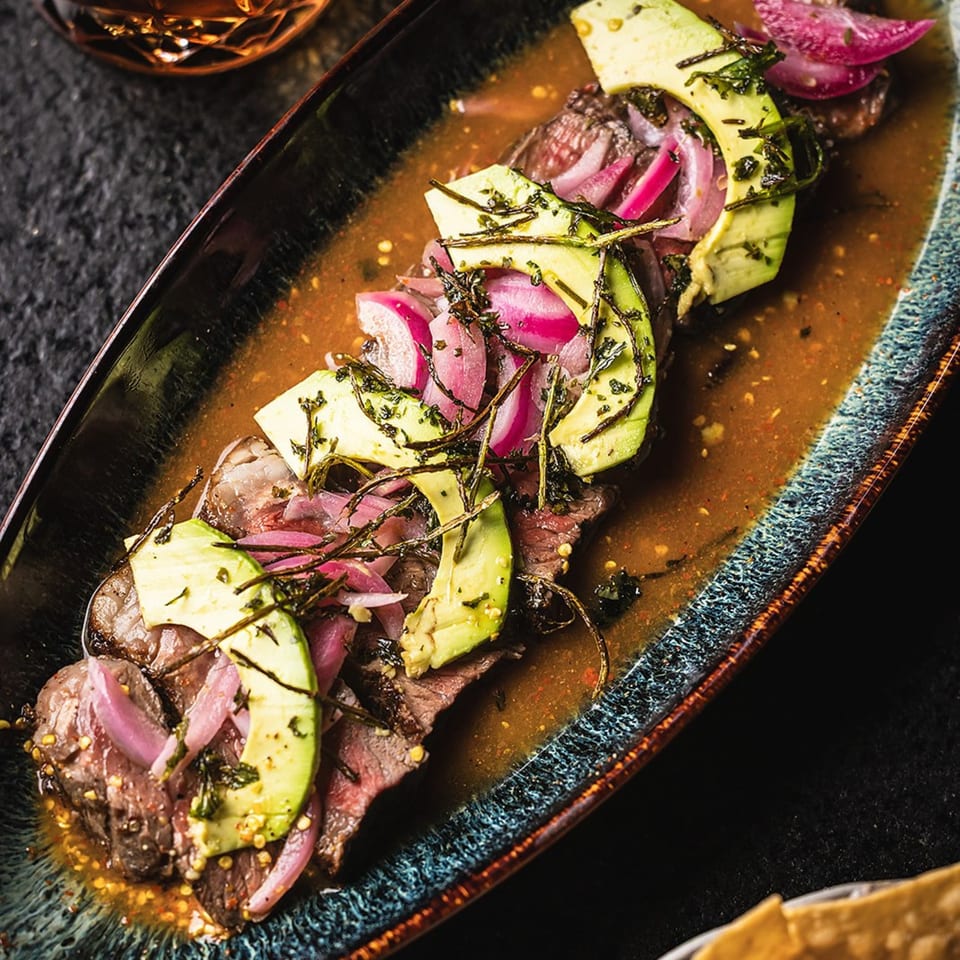 Sliced steak with avocado, pickled red onions, and herbs in a savory sauce on a dark platter.